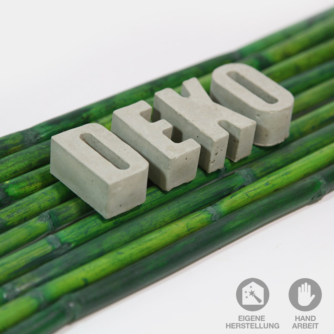 Das Wort "DEKO" aus handgefertigten Beton-Buchstaben liegend auf grünen Bambusstäben.