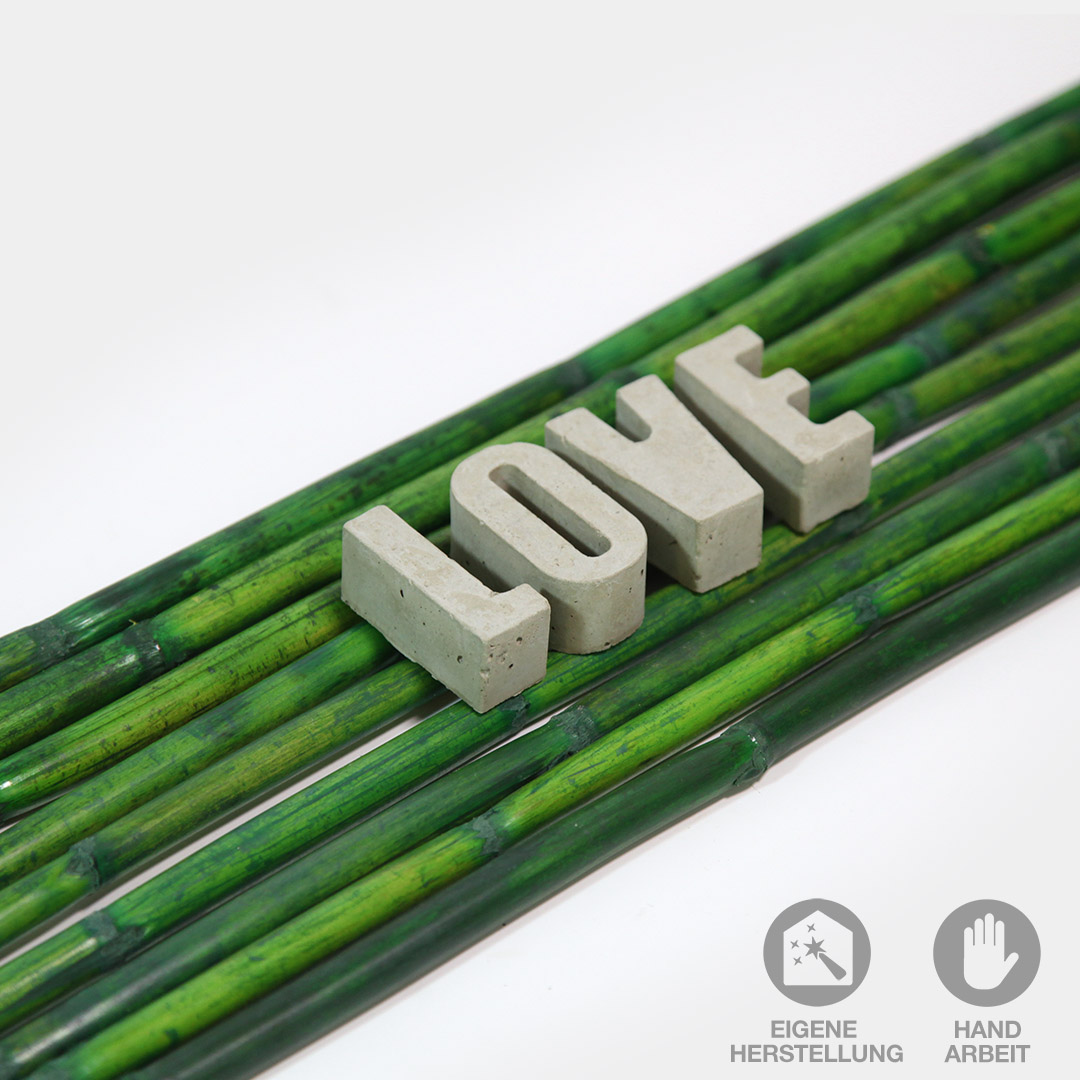 Das Wort "LOVE" aus handgefertigten Beton-Buchstaben liegend auf grünen Bambusstäben.