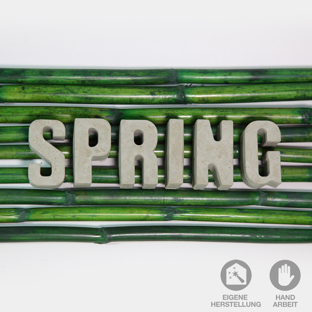 Das Wort "SPRING" für Frühling, aus handgefertigten Beton-Buchstaben liegend auf grünen Bambusstäben.