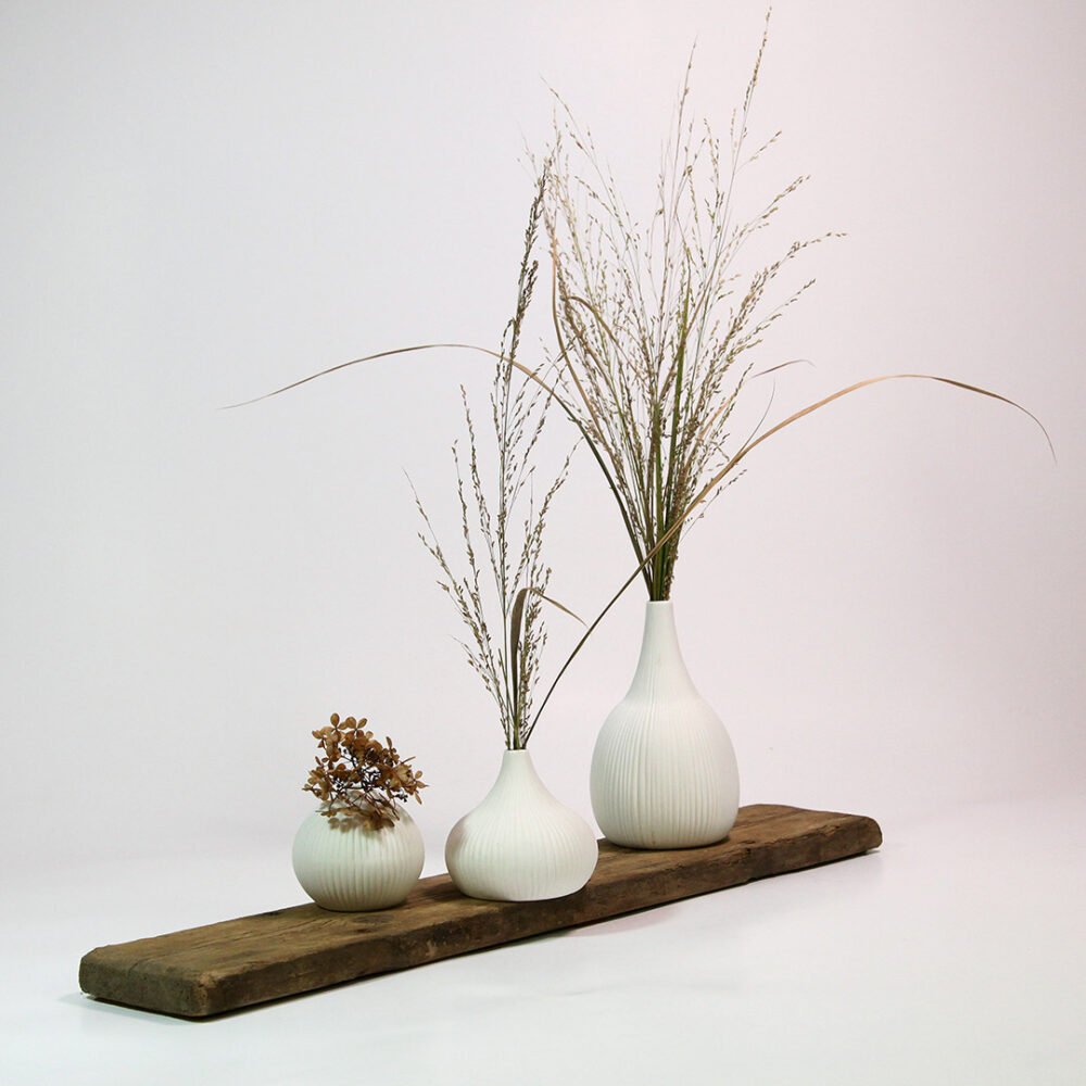 Drei weiße Keramik-Vasen im Scandi-Style bestückt mit Trockenblumen und Gräsern auf einer alten Holzplanke.