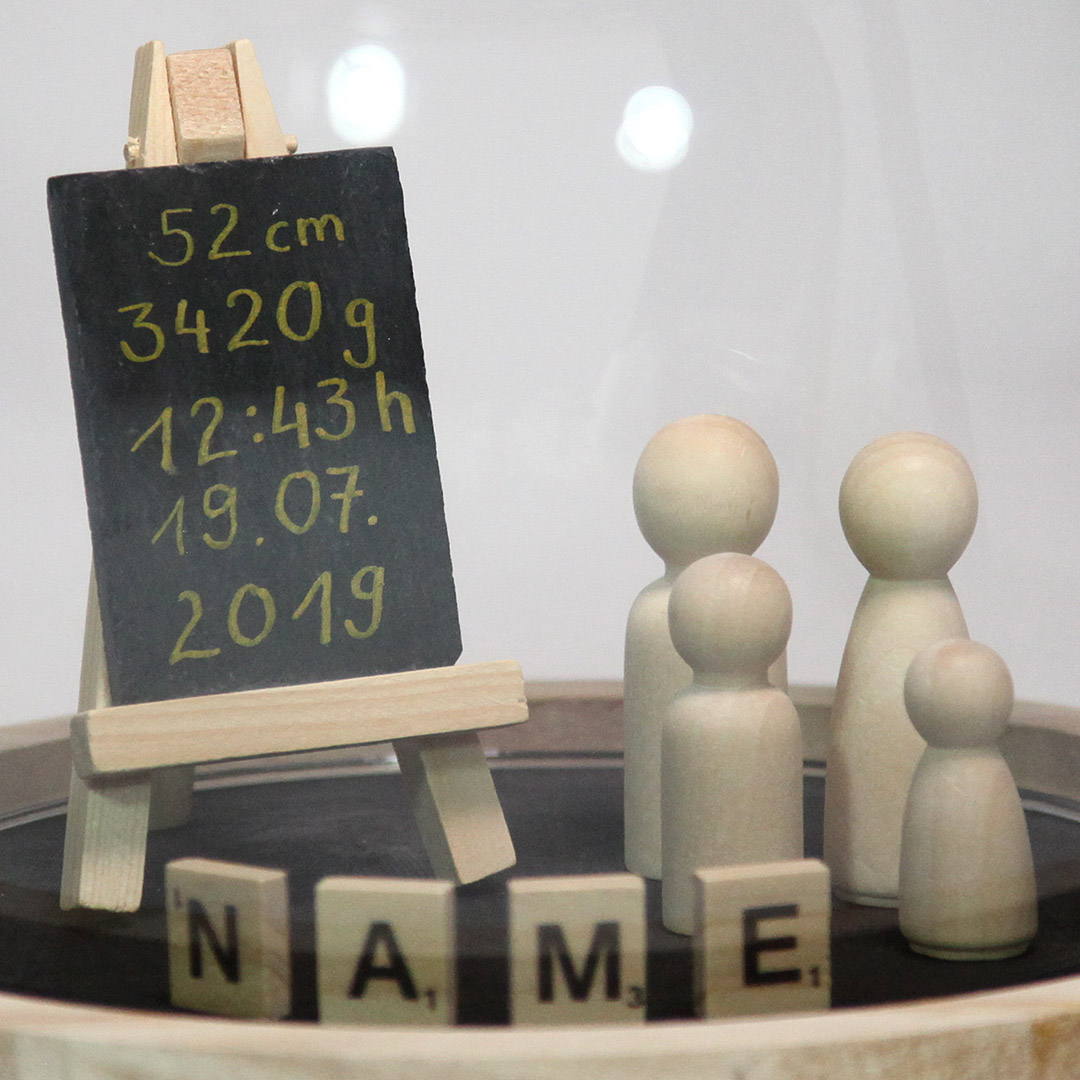 Ein Ausschnitt unserer schlichten, dekorativen Glas-Haube. Bestückt mit Holz-Figuren, Namens-Blättchen und einer mit den Geburtsdaten beschrifteten Schiefern-Tafel.