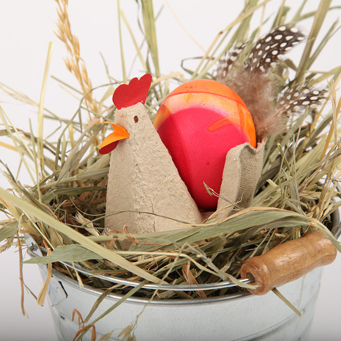 Osterdeko Huhn Mathilde im Eimer. Ein in Handarbeit gefertigtes Papier-Huhn namens Mathilde sitzt weich gebettet im Heu eines Zink-Eimers.