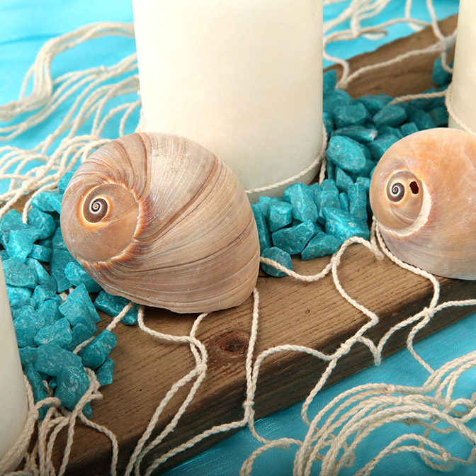 Nahaufnahme von unserem dekorativem Holz-Brett mit Muscheln, türkisfarbenen Deko-Steinen , Netz und wollweißen Kerzen.